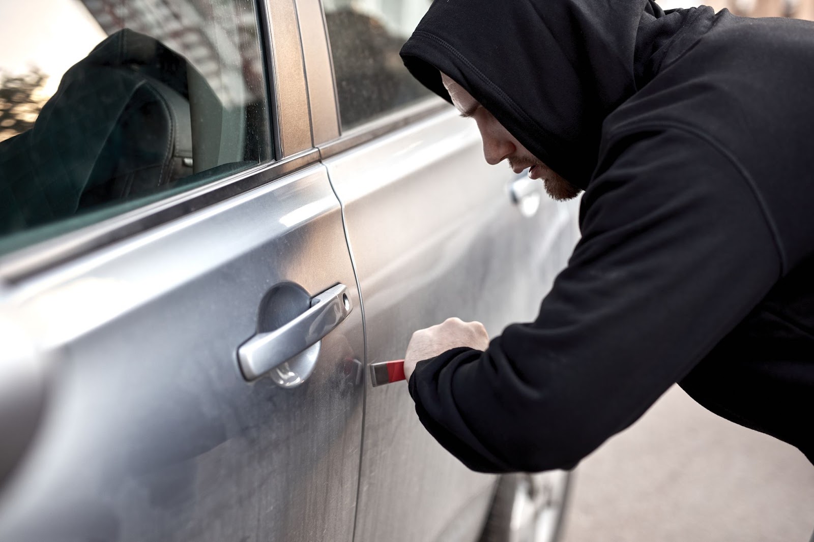 Zlodej v čiernej mikine s páčidlom, snažiaci sa ukradnúť strieborné auto cez predné dvere.