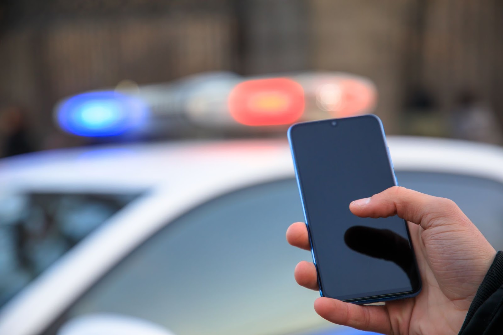 Ruka držiaca telefón pred policajným autom so zapnutými majákmi.